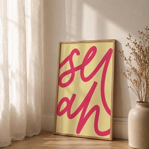 Könnte beinhalten: Ein rechteckiges gerahmtes Wandbild mit hellgelbem Hintergrund und dem Wort "yeah" in einer großen, fetten, kursiven pinkfarbenen Schrift. Der Rahmen besteht aus hellem Holz und hält den Druck sicher innerhalb seiner Ränder.