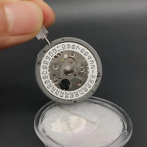 K&ouml;nnte beinhalten: Nahaufnahme eines Uhrwerks mit Datumsrad. Das Uhrwerk ist grau mit silbernen Komponenten und einem wei&szlig;en Datumsrad mit schwarzen Zahlen. Ein kleines Werkzeug wird &uuml;ber dem Uhrwerk gehalten.
