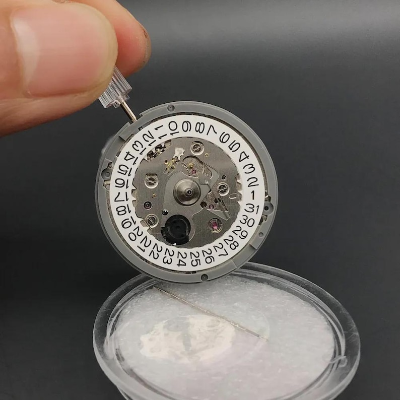K&ouml;nnte beinhalten: Nahaufnahme eines Uhrwerks mit Datumsrad. Das Uhrwerk ist grau mit silbernen Komponenten und einem wei&szlig;en Datumsrad mit schwarzen Zahlen. Ein kleines Werkzeug wird &uuml;ber dem Uhrwerk gehalten.