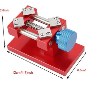 Pode incluir: Ferramenta de reparo de relógio de metal vermelho com uma maçaneta azul e quatro grampos de metal prateados. A ferramenta mede 16,5 cm por 12 cm por 6,5 cm.