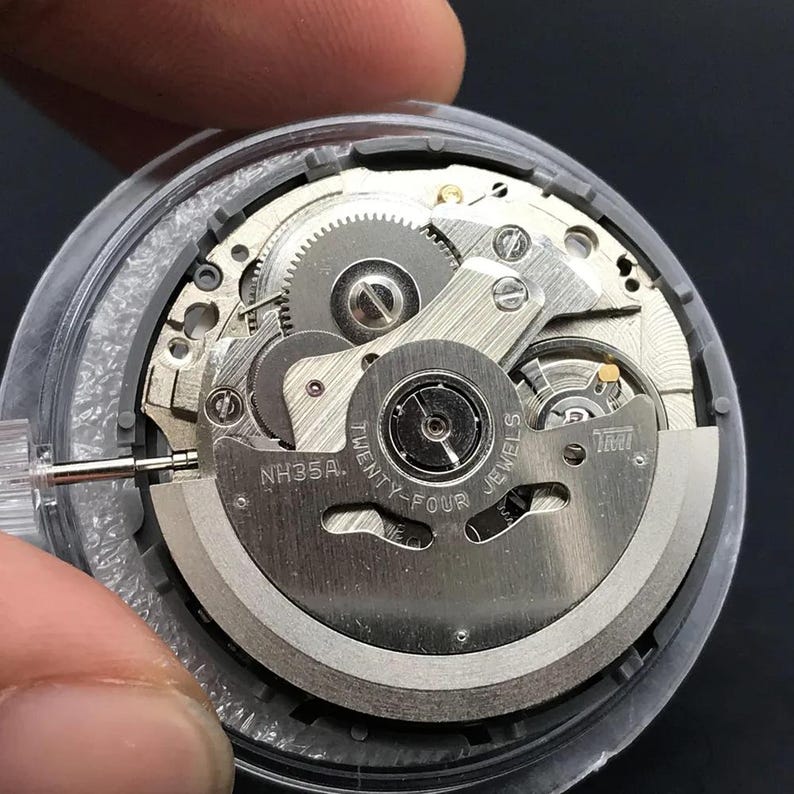 K&ouml;nnte beinhalten: Nahaufnahme eines Uhrwerks mit filigranen Zahnr&auml;dern und Komponenten. Der silberfarbene Mechanismus ist mit "NH35A" und "Twenty-Four Jewels" beschriftet. Das Uhrwerk befindet sich in einem transparenten Geh&auml;use, das die inneren Funktionen der Uhr zeigt.