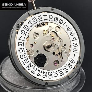 Pode incluir: Uma visão detalhada do movimento do relógio Seiko NH35A. O mostrador prateado exibe a data com algarismos pretos. O texto "SEIKO NH35A Metal movement made in Japan" está presente. O intrincado mecanismo do relógio é visível.