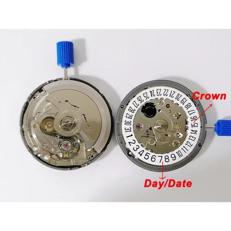 K&ouml;nnte beinhalten: Zwei Uhrwerke mit sichtbaren Zahnr&auml;dern und Komponenten. Eines hat ein wei&szlig;es Zifferblatt mit den Zahlen 1-31, beschriftet mit "Day/Date" und "Crown". Beide haben blaue Einstellstifte. Die Uhrenteile liegen auf einer wei&szlig;en Oberfl&auml;che.