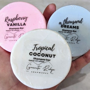 May include: Three round shampoo bars, one pink, one white, and one light blue. Each bar has a different scent: Raspberry Vanilla, Tropical Coconut, and A Thousand Dreams. Each bar is labeled with the name of the scent, "Shampoo Bar", the approximate weight, and "Granite Ridge Soapworks".