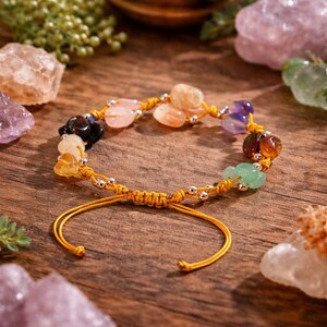 May include: A handmade bracelet featuring a variety of colorful gemstones, including amethyst, citrine, and rose quartz. The stones are strung on a yellow cord with silver-colored beads. The bracelet is displayed on a wooden surface, showcasing its natural beauty.
