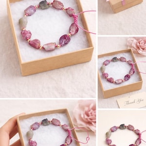 May include: A gemstone bracelet featuring a series of irregularly shaped, pink and purple tourmaline stones, interspersed with small gold-colored beads. The bracelet is presented in a small, square, brown gift box with a white interior.