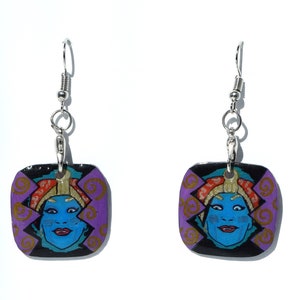 Könnte beinhalten: Ein Paar quadratische Ohrringe mit einem blauen Gesicht, roten Lippen und einer goldenen Kopfbedeckung. Das Gesicht ist vor einem lila und schwarzen Hintergrund mit goldenen Akzenten platziert. Die Ohrringe hängen an silberfarbenen Ohrhaken.