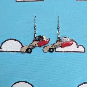 May include: A pair of dangle earrings featuring a graphic illustration of a vintage-style cream-colored car with red accents and a dark-haired driver, suspended from silver-toned metal hooks. The design shows a side profile of the vehicle.