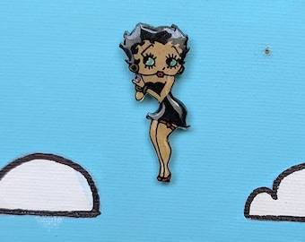 Op de afbeelding: Een kleine emaille speld met een stripfiguur met zwart haar, grote ogen en een zwarte jurk, geplaatst tegen een lichtblauwe achtergrond met witte wolken. Het figuur heeft een getinte huid, rode lippen en rode details aan de kousenbanden.