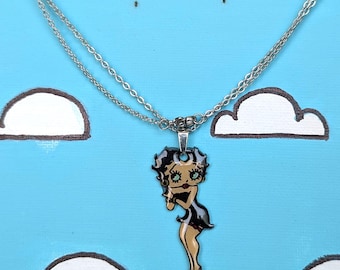 May include: A silver-toned metal chain necklace featuring a small, flat pendant of a cartoon character with black hair, a black dress, and red shoes. The pendant is suspended from a delicate double-strand chain with a metallic finish and a clasp.