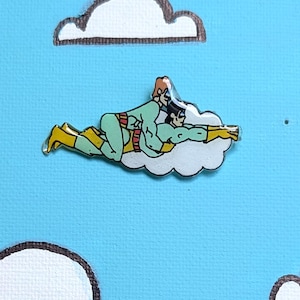 May include: A glossy pin featuring two figures in light green superhero costumes with yellow boots and belts, flying together on a white cloud. The design is set against a light blue background with additional white cloud shapes.