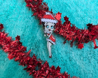 Op de afbeelding: Een decoratieve acrylspeld met een stripfiguur met zwart haar, grote ogen en rode lippen, gekleed in een rood-witte kerstmuts en een bijpassende rood-witte jurk met witte rand, geplaatst tegen een achtergrond van rode kerstslingers.