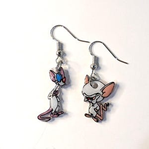 May include: A pair of silver earrings featuring cartoon characters from the animated series "Pinky and the Brain". The earrings are dangle style with a silver hook.