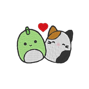 Op de afbeelding: Borduurwerk van een groene dinosaurus en een driekleurige kat met een rood hart ertussen. De dinosaurus en de kat glimlachen en hebben hun ogen dicht.