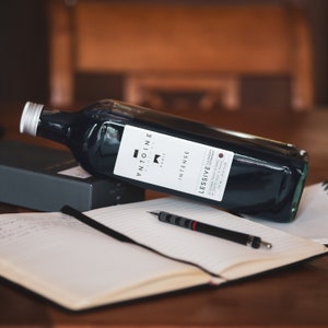 May include: A dark blue glass bottle with a white label that reads "L'ETOINE PARIS INTENSE LESSIVE LAUNDRY DETERGENT" and a red dot. The bottle is lying on a notebook with a pen.