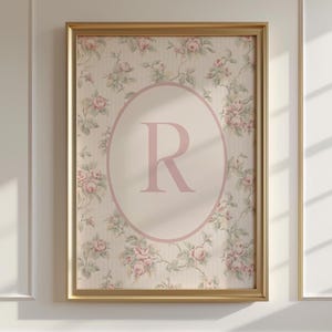 May include: A framed floral print with a gold frame. The artwork features a cream background with a pink oval in the centre, displaying the letter "R" in a soft pink hue. The background is decorated with a delicate rose pattern.