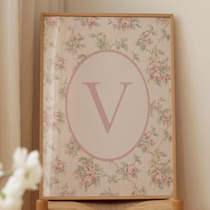 May include: A framed floral print featuring a large letter "V" in a light pink oval. The background has a vintage floral pattern with pink roses and green leaves on a cream-colored striped background. The frame is made of light brown wood.