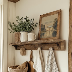 May include: A distressed wooden shelf holds a framed landscape print, white jugs, and a wicker basket with foliage. Below, a woven scrub, wooden chopping board, and hanging tea towels enhance the rustic style.