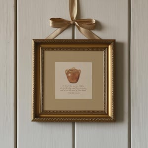 May include: A framed print with a gold frame and a watercolour illustration of a brown pottery jar. The frame is suspended by a gold ribbon. The print includes text from Isaiah 64:8. The frame is square in shape.