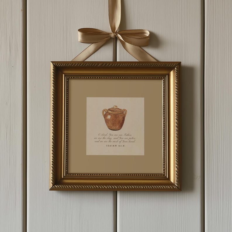 May include: A framed print with a gold frame and a watercolour illustration of a brown pottery jar. The frame is suspended by a gold ribbon. The print includes text from Isaiah 64:8. The frame is square in shape.