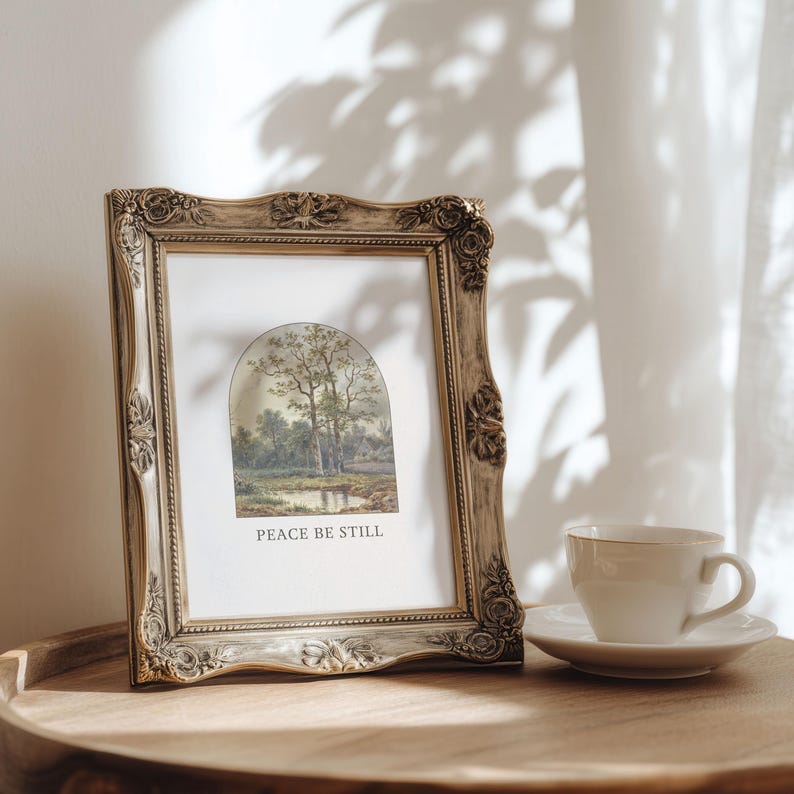 May include: An antique-style picture frame with a landscape print and the text "PEACE BE STILL." The frame is light brown with decorative detailing. A white teacup and saucer are on a wooden surface.