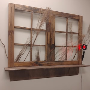 Puede incluir: Un marco de ventana de madera marrón oscuro con un estante a juego fijado en la parte inferior. El marco presenta varios paneles rectangulares y está decorado con ramas finas y secas. Se ha fijado una pequeña luz negra con brillo rojo.