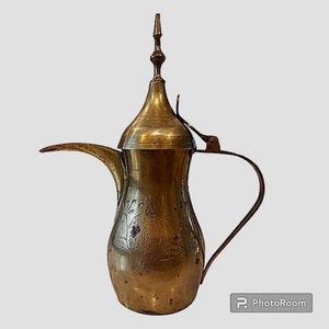 May include: A traditional brass Arabic coffee pot with an ornate design. The pot has a spout and a handle, and a lid with a finial.