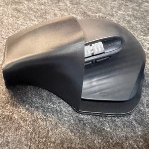 May include: A black ergonomic computer mouse featuring a contoured shape, a prominent thumb rest, and a metallic silver scroll wheel positioned on the side. The device has a matte finish and includes additional side buttons located near the scroll wheel.