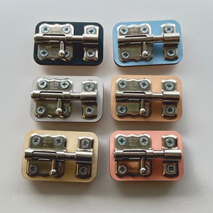 Puede incluir: Seis cerrojos de puerta mostrados en diferentes fondos de colores. Los cerrojos son plateados con una manija negra y están montados en fondos negro, azul, blanco, beige, marrón claro y rosa.