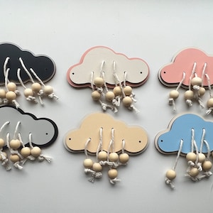 May include: Six wooden cloud-shaped toys in various colours: black, grey, white with pink trim, pink, yellow, and blue. Each cloud has wooden beads attached with white string, resembling raindrops. A fun, educational toy for children.