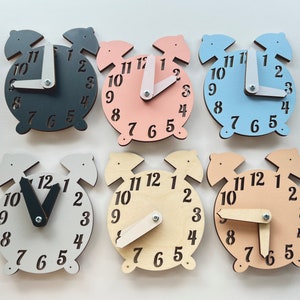 May include: Six wooden alarm clocks in different colors, including black, pink, light blue, white, beige, and peach. Each clock has a movable hour and minute hand.