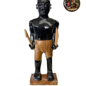 May include: A carved wooden statue of a figure with black skin, wearing brown shorts and holding wooden tools. The statue is on a wooden base. A circular emblem with text and a tiger is in the upper right corner.