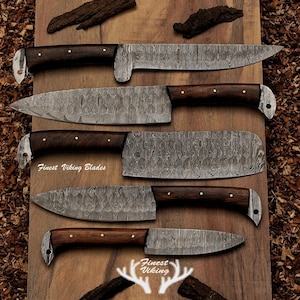 May include: Five Damascus steel kitchen knives with wooden handles on a wooden surface. The knives have a distinctive pattern and are labelled "Finest Viking Blades".