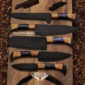 May include: Set of five black kitchen knives with wooden handles featuring a blue and gold inlay. The knives are arranged on a wooden surface with wood shavings. The text "Finest Viking Blades" is visible on the surface.