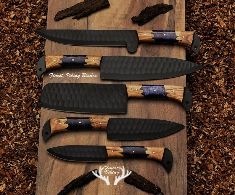 May include: Set of five black kitchen knives with wooden handles featuring a blue and gold inlay. The knives are arranged on a wooden surface with wood shavings. The text "Finest Viking Blades" is visible on the surface.