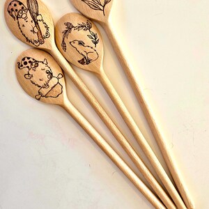 May include: Five wooden spoons with engraved designs. Four spoons feature whimsical illustrations of gnomes with cooking tools and floral or botanical elements. The spoons are made of light-colored wood.