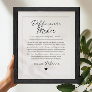 May include: A black framed print with a white background and black text. The text defines the word "Difference Maker" and describes the qualities of a person who makes a difference in the world. The print includes the name "Rebecca" at the bottom.