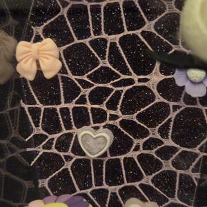 May include: A decorative display featuring a black background with a pink web-like pattern. Adorned with pastel-colored bows, heart-shaped and flower-shaped decorations, and artificial flowers. A small purple couch with a cartoon character is also present.