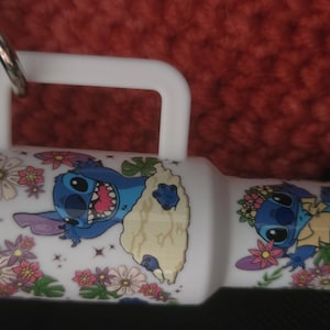 May include: White tumbler with a handle, featuring a cartoon Stitch design with floral accents. The tumbler has a silver ring and a white handle. The design includes Stitch with a flower crown and Stitch with a happy expression.