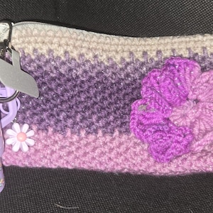 May include: A crocheted, ombre-colored pouch in shades of purple and pink, adorned with a large, three-dimensional purple flower. A small, decorative daisy and a Winnie the Pooh themed tumbler with a straw are also present.