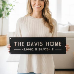 May include: A black metal sign with the text "THE DAVIS HOME" in white, and coordinates below. A person holds the sign in a bright living room. The sign is a decorative home accessory.
