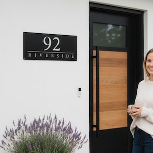 May include: A black metal address sign with the number 92 and the word "Riverside" in white lettering. The sign is mounted on a white wall next to a wooden door. A woman is standing in front of the door.