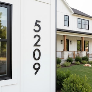 Könnte beinhalten: Schwarze Hausnummern 5209 an einer weißen Außenwand angebracht. Die Zahlen sind in einer modernen Schriftart gehalten. Im Hintergrund befindet sich ein Haus mit weißer Fassade und schwarzen Zierleisten. Grüne Büsche und Gras befinden sich im Vordergrund.