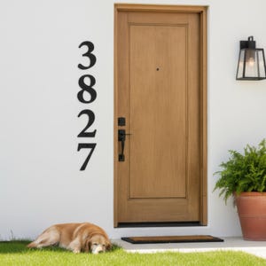 Vertical black metal house number sign mounted on white wall beside wooden front door, modern outdoor address plaque