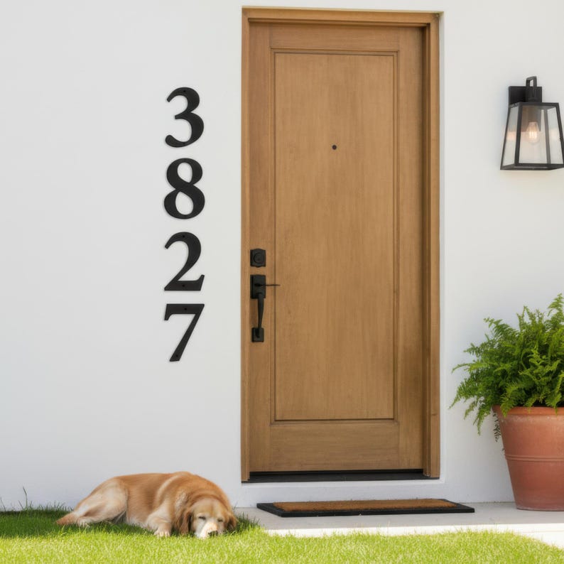 Vertical black metal house number sign mounted on white wall beside wooden front door, modern outdoor address plaque