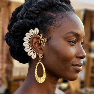 May include: A gold-toned wire ear cuff featuring a curved row of white cowrie shells along the outer edge. The piece includes a decorative wire swirl at the top and a dangling, hammered gold-toned oval pendant suspended from the lower section.