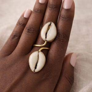 Op de afbeelding: Een goudkleurige ring met twee ovale, witte kaurischelpen. De schelpen hebben een zwart en bruin interieur en zijn gezet in goudkleurig metaal. De ring zit aan een vinger.