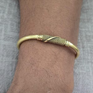 May include: A gold-colored cuff bracelet. The bracelet has a woven design in the center and is curved to fit the wrist. The metal has a polished finish. The bracelet is a piece of jewelry.