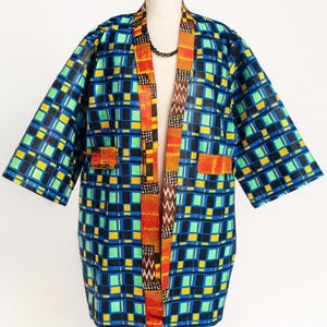 Könnte beinhalten: Eine leuchtende Kimono-Jacke mit offenem Schnitt. Die Jacke hat ein auffälliges geometrisches Muster aus blauen, schwarzen, grünen und gelben Quadraten. Die Verzierungen und Taschen sind mit orangefarbenem und braun gemustertem Stoff versehen. Eine schwarze Halskette ist sichtbar.