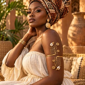 May include: A gold wire arm cuff featuring intricate coiled patterns and two white cowrie shells. The accessory is worn on the upper arm, complemented by a stack of thin gold bangles on the wrist and textured gold disc earrings.